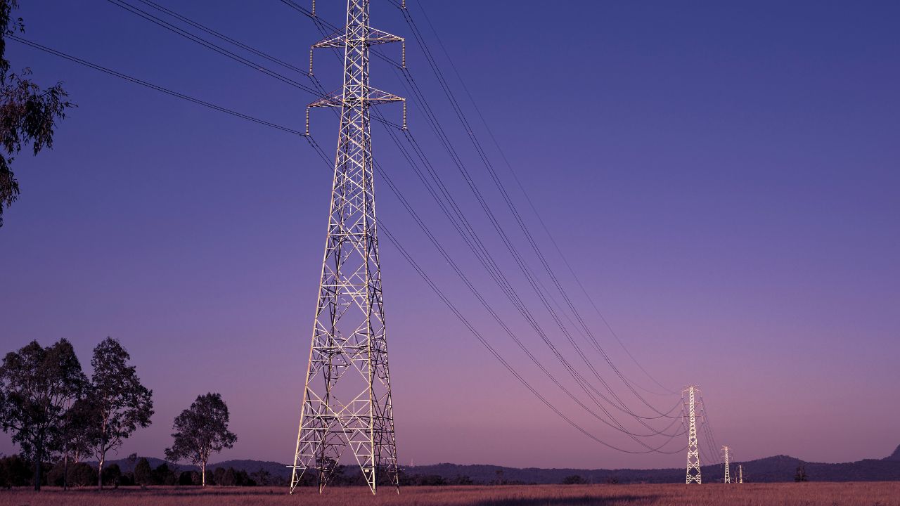 Tall, steel structure carries high voltage power lines across a field with trees and hills in the background