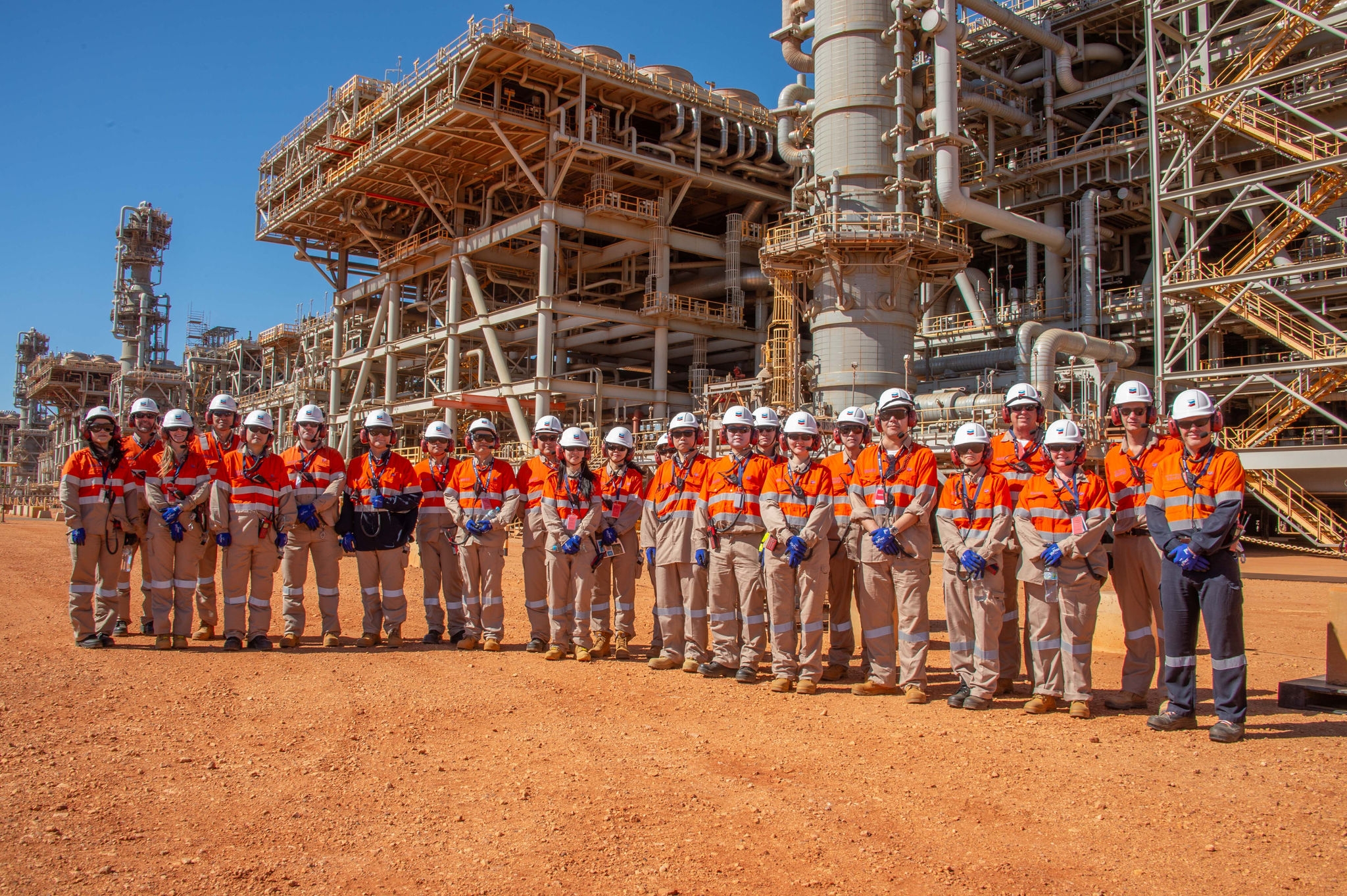 A group of men and women wear orange high visibility vests stand in front of a plant of steel pipes and framework used for carbon capture.