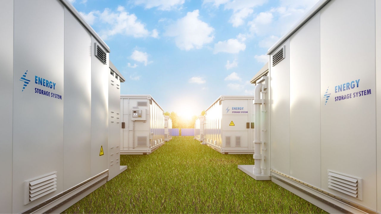 Rectangular, white metal battery storage containers sitting on a grass field with the sun and solar panels in the background