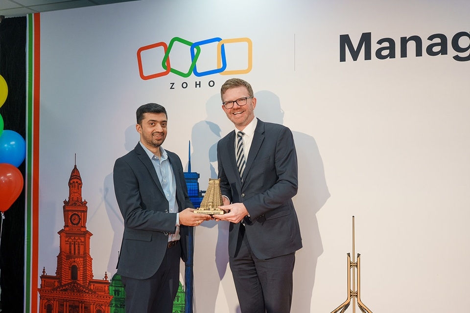 Two businessmen standing together in front of Zoho sign