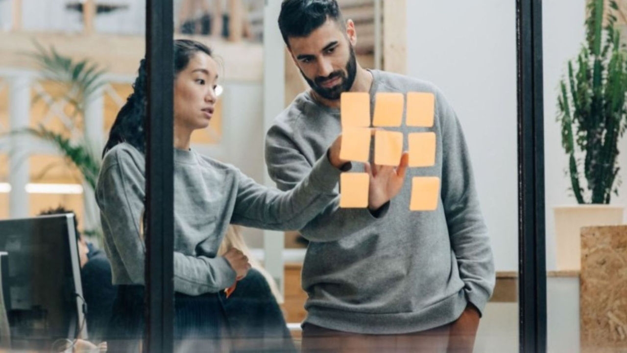 A woman and a man brainstorm using orange sticky note paper squares on a glass wall of a meeting room