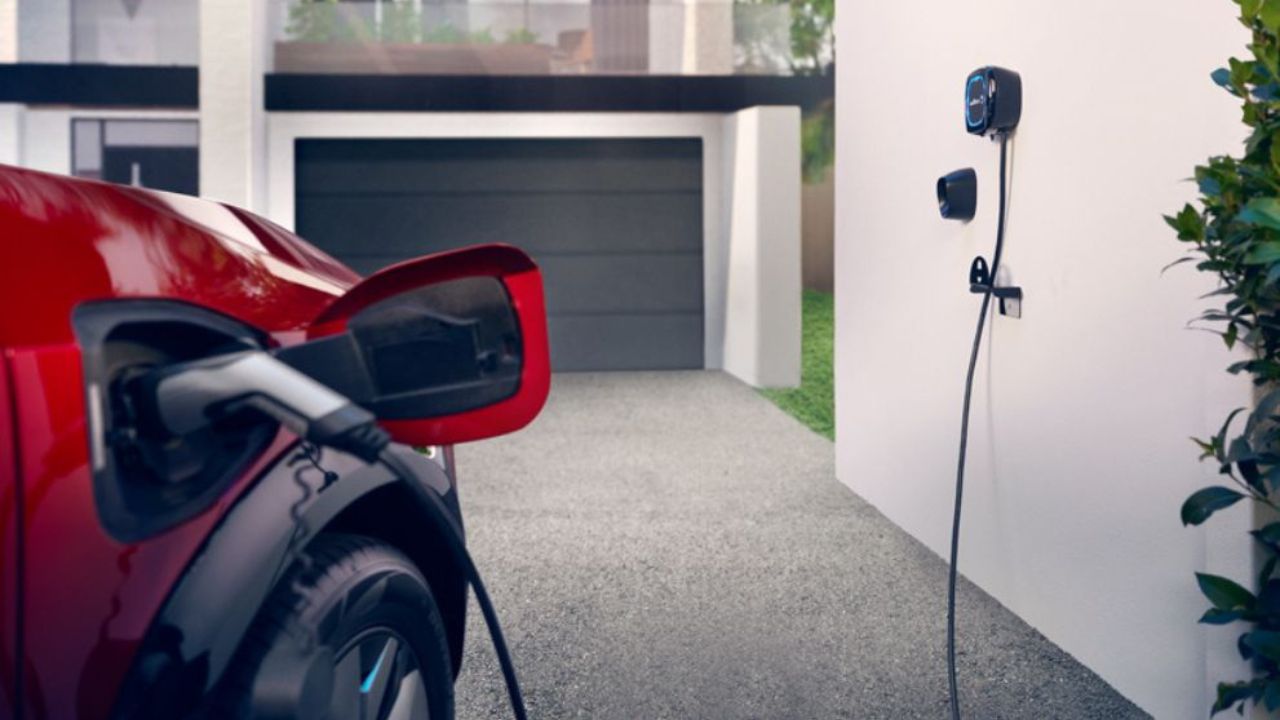 A charging cable from a home charger is connected to a red electric vehicle in a home driveway