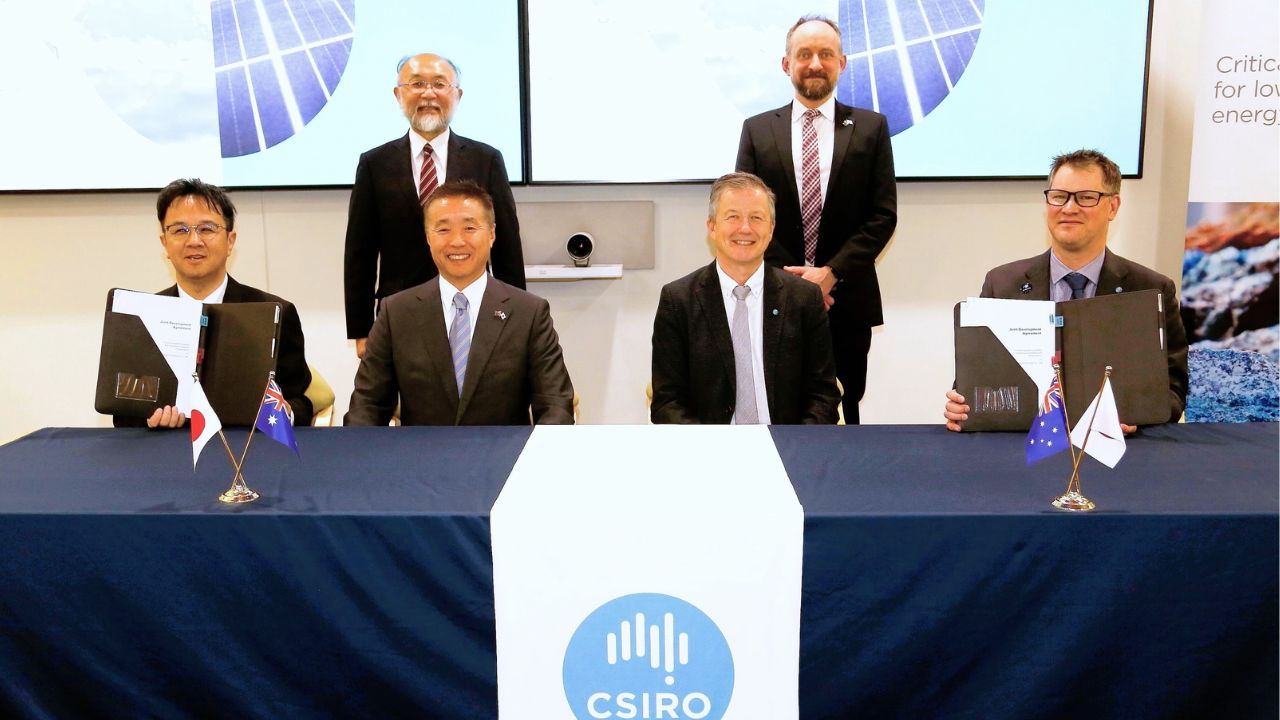 Four business men in suits sit at a table with two holding signed agreement documents. There are two men standing behind them.