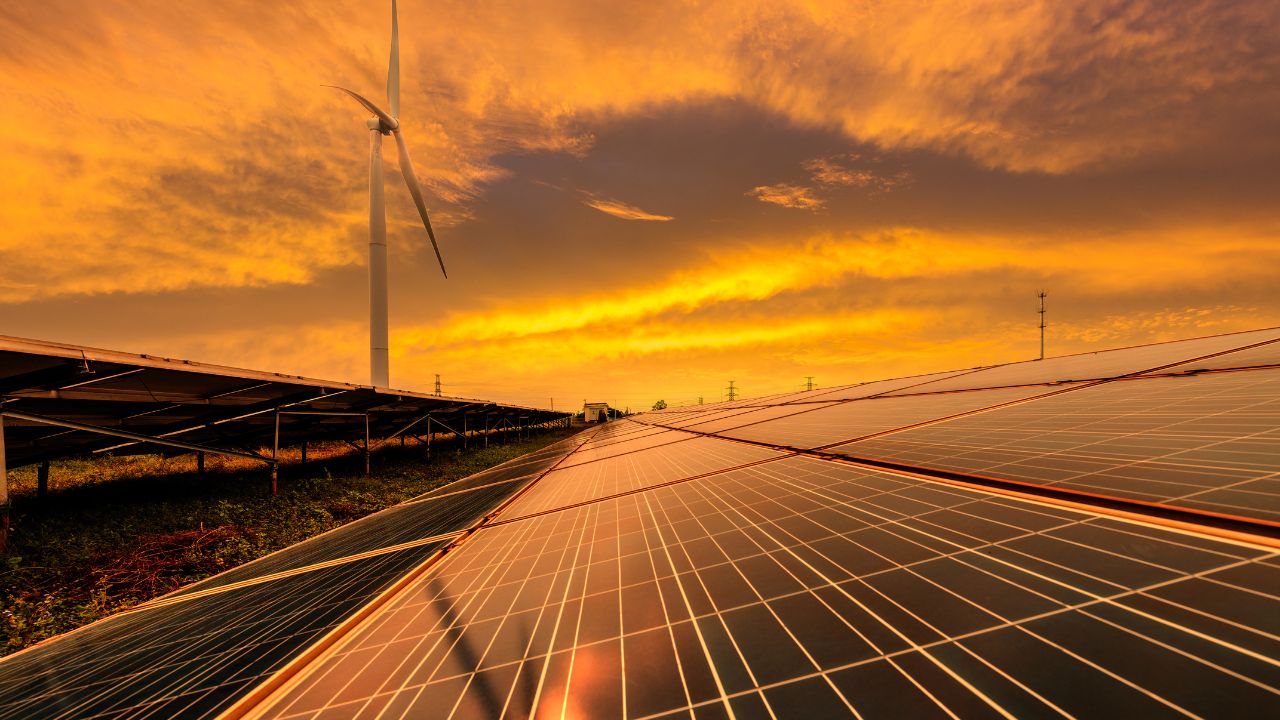 The orange glow of dusk sunlight illuminates banks of solar panels with a wind turbine in the background