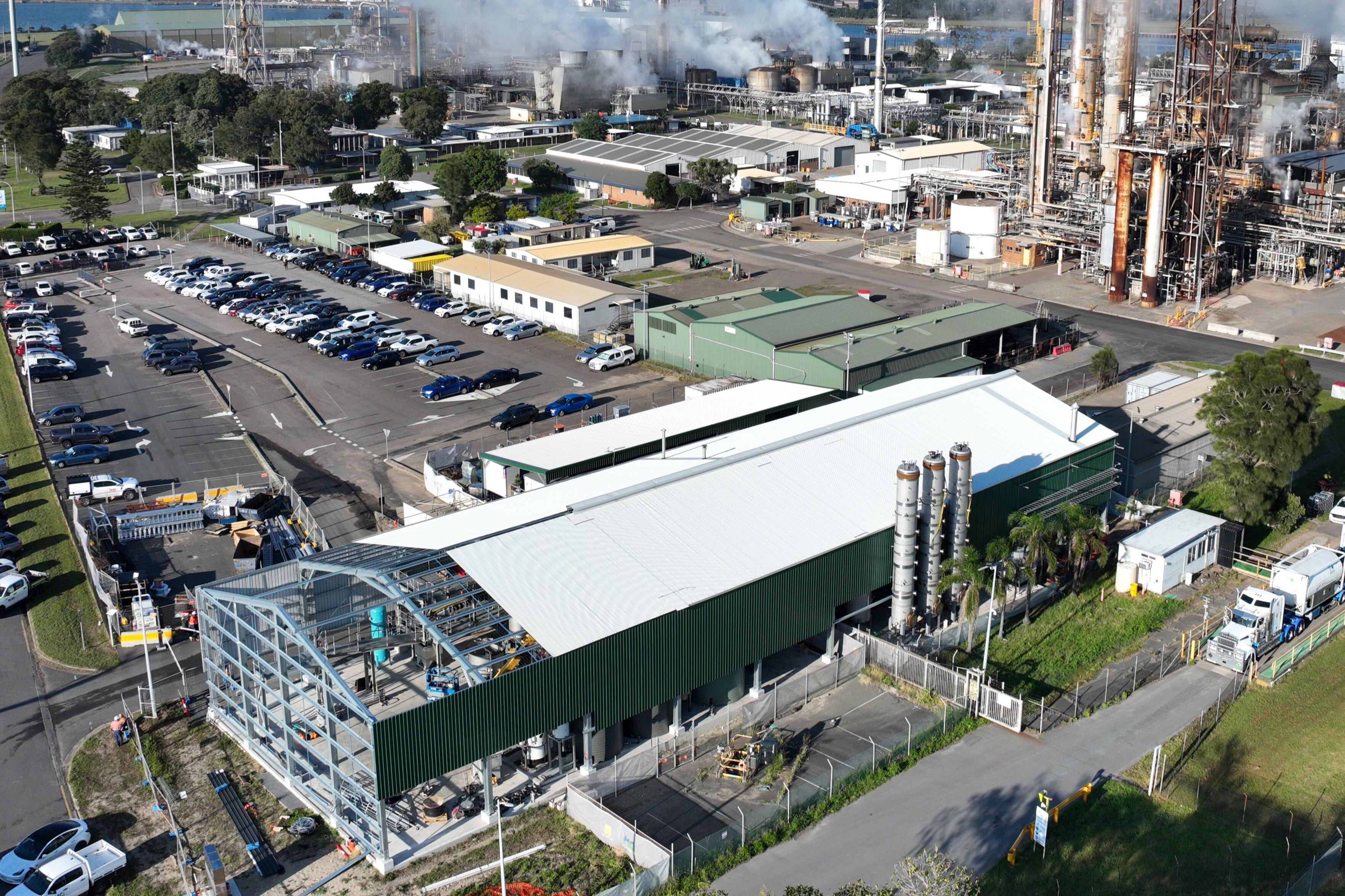 A large rectangular warehouse factory, surrounded by a car park, smaller buildings and other factory works in the background