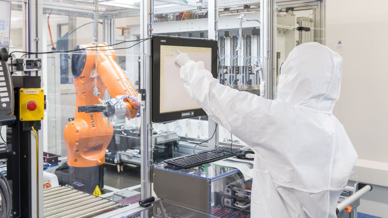 A controller dressed in a white clean suit touches a screen that programs a manufacturing robotic arm