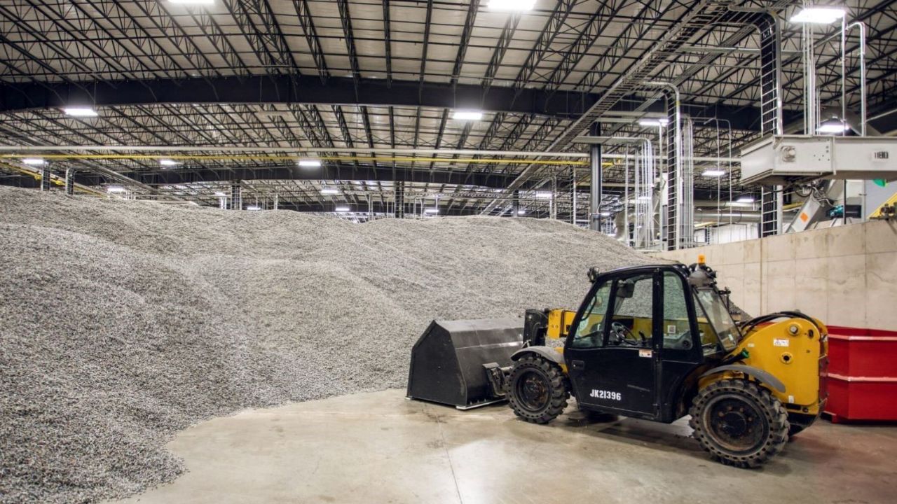 A yellow and black frontend loader is parked inside a large factory building next to a stockpile of recycled plastic.