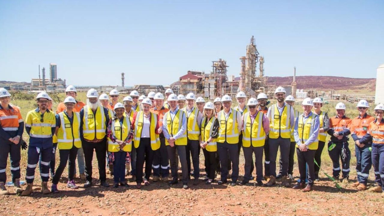 A group of people stand in the Australian outback with a hydrogen processing plant in the background