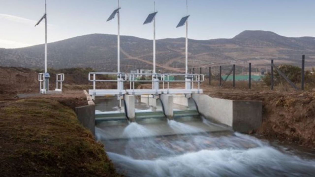 Water flows through three small dam gates with four poles erected above with a single solar panel on each